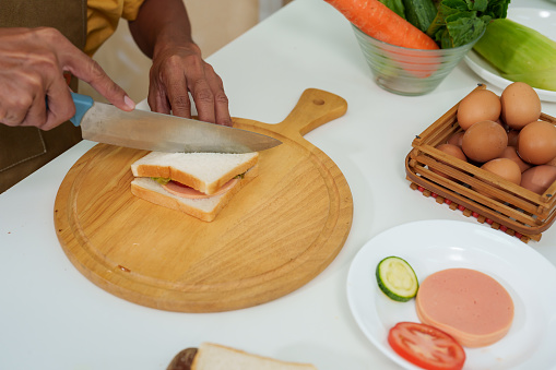 Close-up hands of Asian man is using a knife to cut a sandwich into two halves, making them even. to prepare for breakfast On days when you have to work from home. Wear apron in the kitchen.