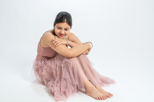 A young woman wearing a pink fit and flare, spaghetti strap net dress, pearl necklace, a golden bracelet on the right hand, pink nail paint on her fingers and toes, hair clipped behind, pink lipstick, sitting and hugging her knees and looking downwards, posing, close-up Studio shot.