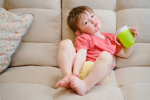 The non-spill cup is in the hands of the smiling child, who sits contentedly on the couch, drinking water. A toddler baby drinks liquid from a green plastic cup. Kid aged about two years