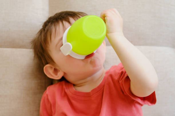 o bebê segura alegremente uma caneca cheia de água, bebendo o líquido branco enquanto descansa no sofá. bebendo de um copo, a criança sorri no sofá. criança com cerca de dois anos de idade - water child bottle little boys - fotografias e filmes do acervo