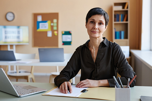 Portrait of mature teacher looking at camera while sitting at her workplace with laptop in class
