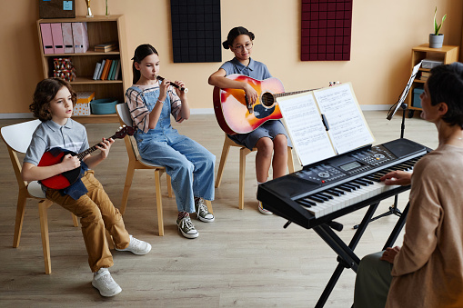 Group of children sitting on chairs and playing musical instruments with teacher playing piano during lesson in music class
