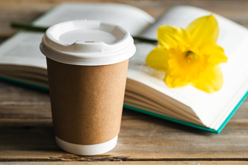 Paper disposable cupfor a drink, a book and a yellow narcissus flower on a blurred wooden background.