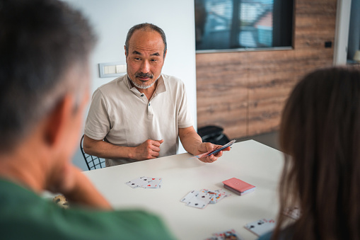 Cheerful Asian Grandfather sitting at a table in a domestic dining room with son and granddaughter. They are playing a card game.
