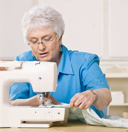 A senior woman sits and uses a sewing machine. She is looking down and concentrating on her work. Square shot.