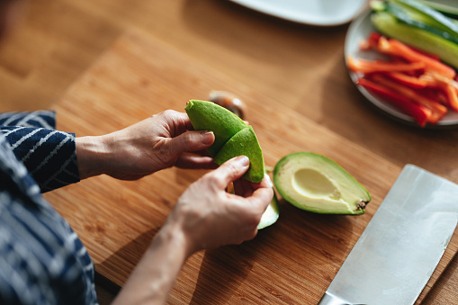 Close up shot of a woman holding cut pieces of avocado. There are cut vegetables in the background.