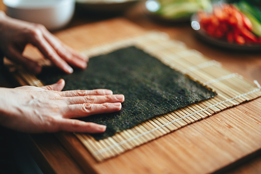 Close up of an unrecognizable woman placing seaweed on a bamboo sushi rolling mat on the wooden cutting board ready to make some sushi rolls.