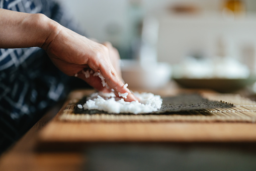 Close up shot of an unrecognizable woman's hands putting rice on a seaweed sheet while making sushi at home.