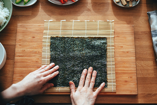 Close up of an unrecognizable woman placing seaweed on a bamboo sushi rolling mat on the wooden cutting board ready to make some sushi rolls.