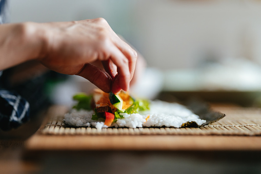 Close up of an unrecognizable woman rolling an open sushi roll with a bamboo rolling mat. She is making a vegetarian roll with tofu, paprika, rice, carrots and lettuce.
