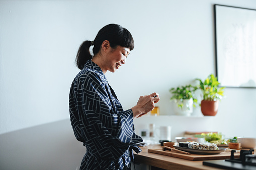 Cheerful Asian woman holding her smart phone and taking a photo of a full plate with sesame seed and seaweed sushi rolls she made at home. She is smiling and looking down.