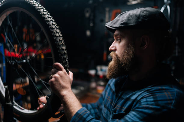 gros plan d’un réparateur barbu utilisant un outil pour tendre des rayons dans la jante d’une roue de vélo travaillant dans un atelier de réparation de vélos avec un intérieur sombre. - cycling bicycle occupation men photos et images de collection