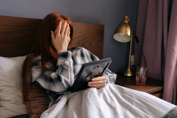 Side view of grief-stricken young woman lying on bed covering face with hand and crying while holding picture frame, touching photograph with love. Side view of grief-stricken young woman lying on bed covering face with hand and crying while holding picture frame, touching photograph with love. Concept of nostalgia, grief, longing and loneliness. person holding frame over face stock pictures, royalty-free photos & images