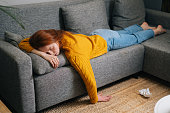 High-angle view of exhausted pretty woman sleeping on cozy couch in living room, lying on stomach. Tired young female with closed eyes resting on sofa