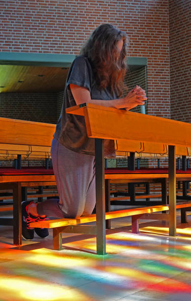 A woman kneeling in prayer in a church. Sunlight streams through the stained glass windows and paints a colorful pattern on the floor