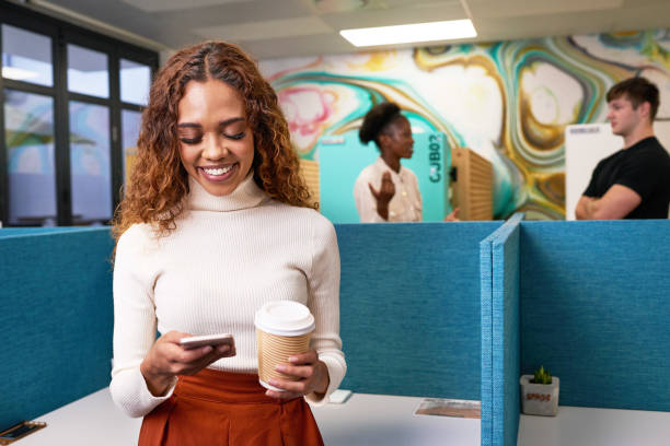 Young multi-ethnic woman texts with cellphone, standing in busy open plan office stock photo