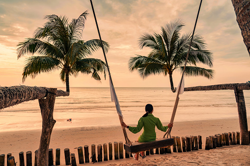 Young asian woman enjoying with wooden swing on the beach in the sunset at tropical island
