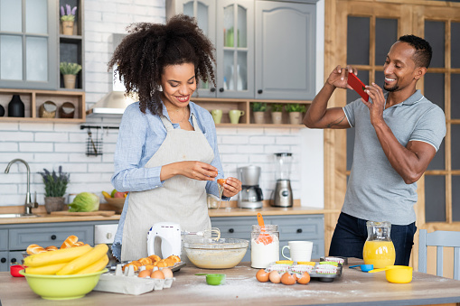 Couple cooking together, having fun time in the kitchen. Multiracial young woman mixing eggs in a bowl, while her boyfriend taking picture of her with his mobile phone, shooting home video or vlogging