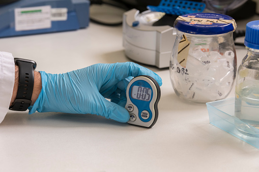 A hand of a lab technician with blue nitril gloves holding an timer in a science laboratory with plastic eppendorfs in glass in the background. Science, analyse, analysing, DNA analysis