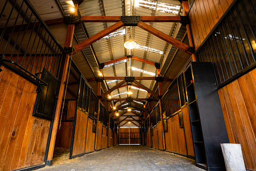 A horizontal low-angle photograph showcasing the interior of rustic wooden horse stables, featuring iron and wood construction elements and a stone floor. The image captures the authentic and traditional architecture of a rural stable.