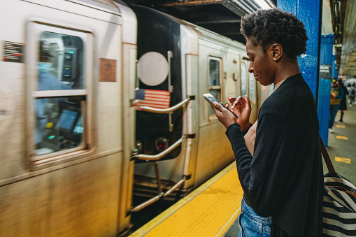 A woman is using a smartphone while she's waiting for the train in a subway station. She's standing in a New York City subway station.
