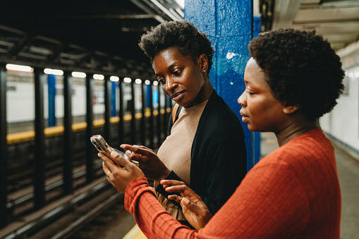 Two friends are using smart phones in a subway station, waiting for the train. They are standing in the subway of New York City.