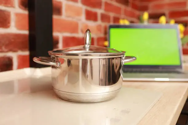 Photo of Steel saucepan on white stylish induction stove and laptop with green screen with chrome key at kitchen on brick wall background. Cooking and vlogging concept