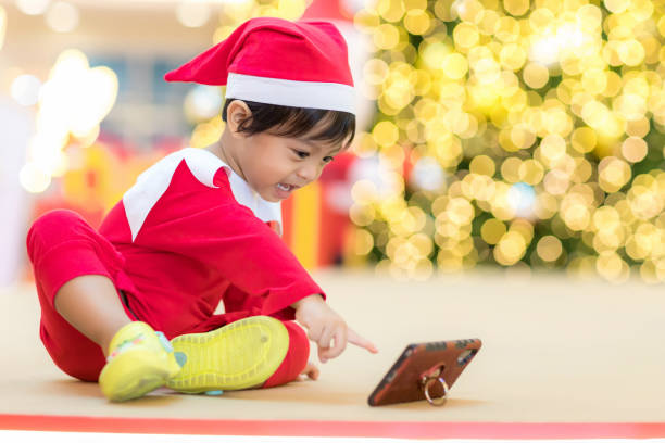 メリークリスマス。赤い帽子をかぶったクリスマスの子供。年末年始。クリスマスのボケ背景にクリスマスの子供たち。 モバイルとソーシャルメディアを使用する子供。彼のスマートフォン - baby holding babies only sign ストックフォトと画像