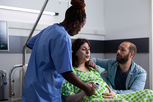 Pregnant woman with painful labor lying in hospital ward bed, having contractions. African american nurse and husband supporting future mother, holding hands for medical assistance