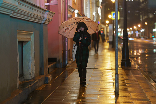 Young Caucasian smiling woman crossing street downtown in rain with umbrella. Autumn time.