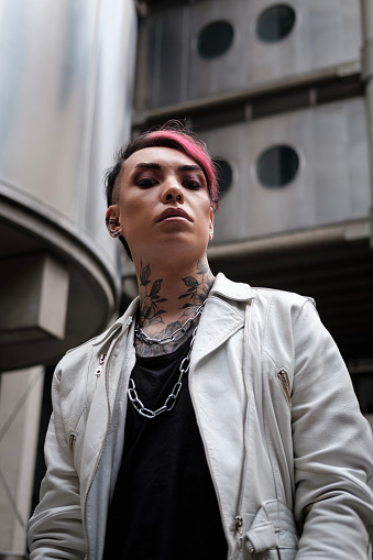 Moody portrait of androgynous non-binary model in futuristic environment in London. He looks so serious, confident and proud. White leather jacket, chains collar and tattoos.