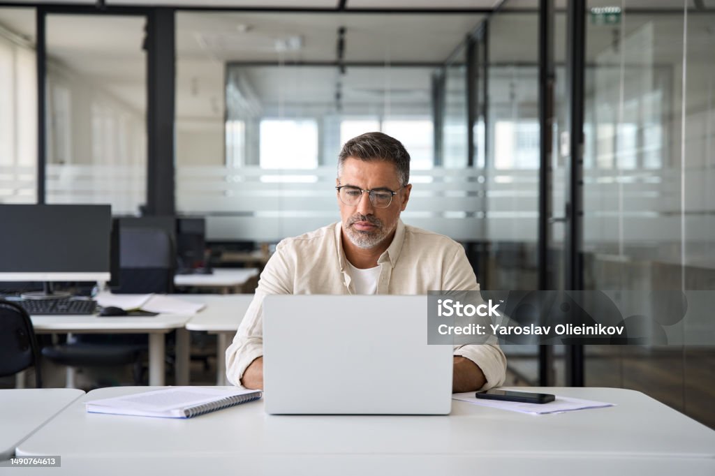 Mature Latin business man ceo trader using computer working in modern office Mature Indian or Latin business man ceo trader using computer, typing, working in modern office, doing online data market analysis, thinking planning tech strategy looking at laptop with copy space. Using Computer Stock Photo