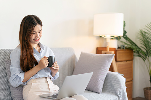 Female freelance is reading business data on laptop and writing information in notebook while sitting on comfortable the couch to drinking coffee and working in living room at home office.