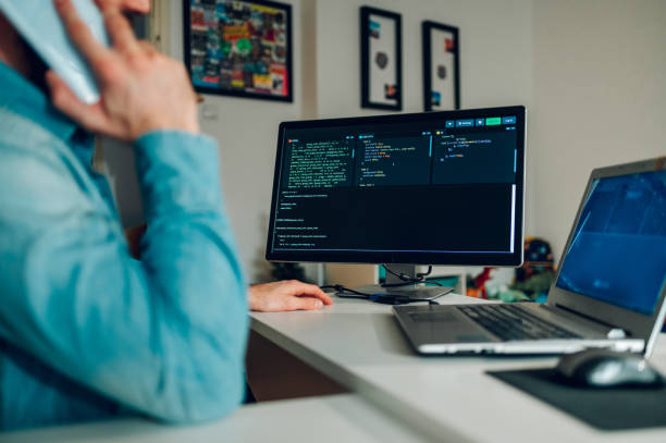 istock Cropped picture of a programmer talking on the phone in a blurry foreground. Selective focus on code on his computer monitor. 1490782074