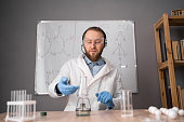 a chemistry teacher in white sits on a chair against the background of a marker board with formulas, conducts an experiment, puts a flask with a chemical on a burner. Webcam view