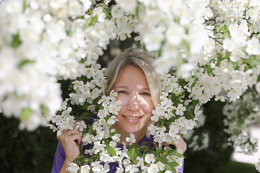 Portrait of a young Caucasian woman standing in the apple tree covered with its' branches blossoming with white flowers in spring. White apple blossom. Blossom season in Ontario, Canada. White apple tree blossom. Mother's Day 2023. Springtime in Canada.