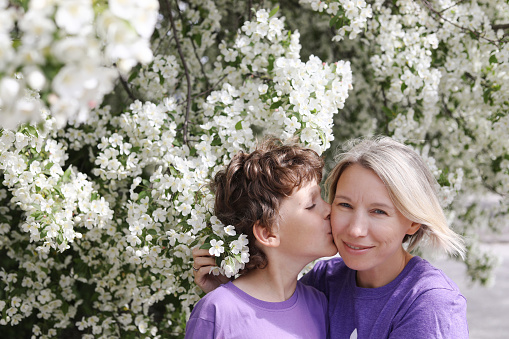 Caucasian boy kissing his mom on the cheek. Mother's Day 2023. Springtime in Canada. Mother and son kissing near the apple tree blossoming with white flowers. Love between mother and son. Celebrating moms.