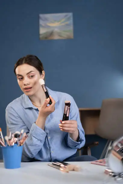 Photo of Happy young blogger applying blush on her face with a brush and looking at the mirror while sitting at the table at home. copy space.