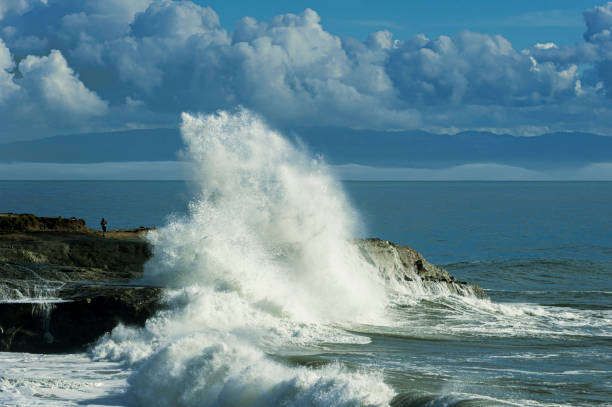 Large powerful Pacific Ocean wave crashing on the rocky shore along the California coast, with an unrecognizable person about to be drenched with water. \n\nTaken in Santa Cruz, California, USA.