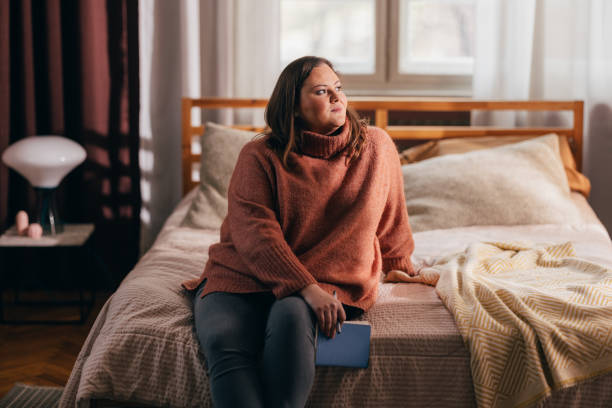 Beautiful Plus Size Woman Sitting on her Bed Looking Away, Holding a Blue Notebook on her Lap