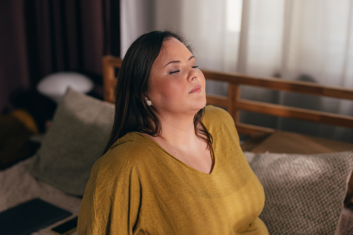 A smiling overweight woman in a yellow T-shirt sitting on her bed trying to relax, taking deep breaths with her eyes closed