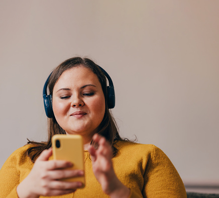 A portrait of a happy overweight woman in a yellow blouse with her headphones on, listening to a podcast on her mobile phone.