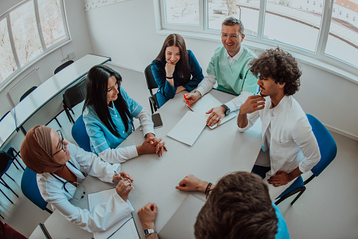 Top view of a group of multiethnic medical professionals including doctors, surgeons, and nurses are gathered in a hospital setting discussing patient care and using modern technology to address challenges in the medical field.