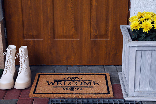 Doormat with word Welcome, stylish boots and beautiful flowers on floor near entrance