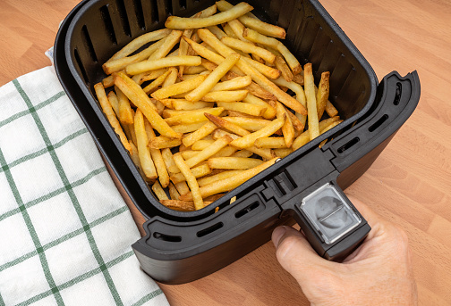 HAND HOLDING AIR FRYER BASKET WITH FRESHLY MADE FRENCH FRIES OR FRIED POTATOES IN THE KITCHEN COUNTERTOP. TOP VIEW.