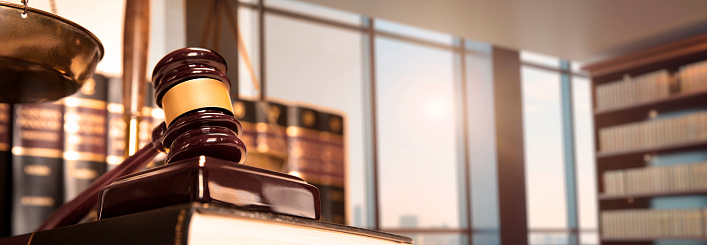 A gavel rests in front of a row of law books in a contemporary law office that looks out toward a big city as the late afternoon sun filters in through the windows.