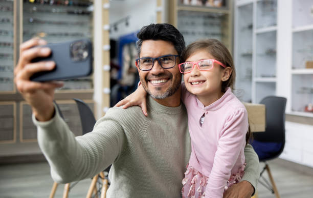 pai e filha tirando selfie experimentando óculos em loja de optometria - optometrist store retail glasses - fotografias e filmes do acervo