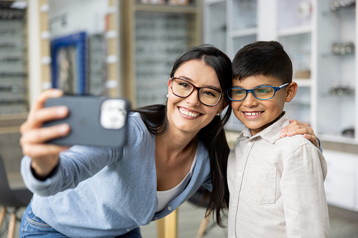 Happy Latin American mother and son taking a selfie trying glasses at an optometry shop and smiling