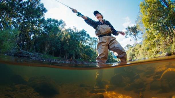 mujer pescadora en el río. la mujer se para en el agua en zancudas y lanza la línea. mujer pescando en el río - fisherwoman fotografías e imágenes de stock
