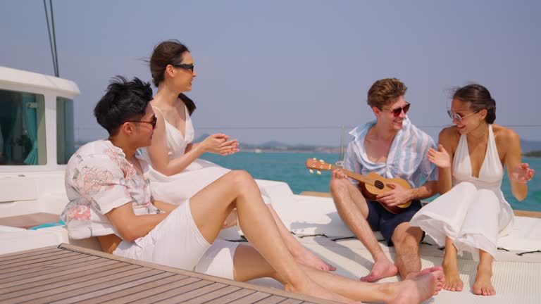 Group of diverse friend sit on deck of yacht while yachting together. Attractive young men and women hanging out, celebrating holiday vacation trip while catamaran boat sailing during summer sunset.